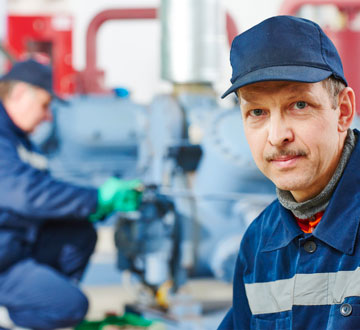 Two workers in blue uniforms; one looks at the camera, while the other works on machinery in the background.