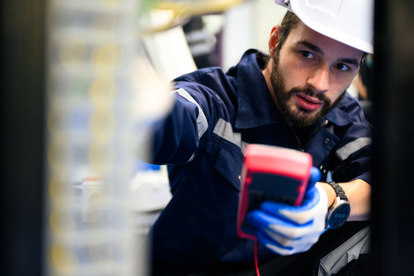 Technician in a helmet and gloves using a handheld device to check electrical equipment in a utility room.