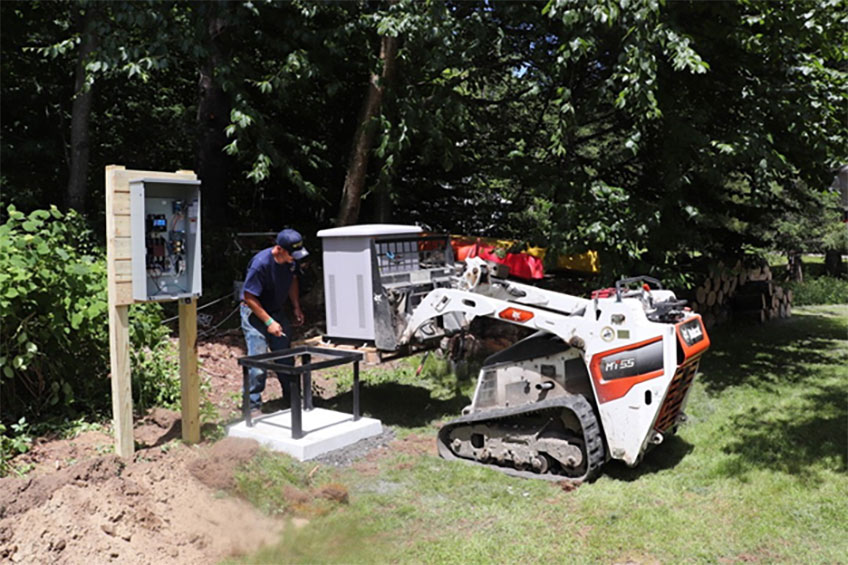 Person operating machinery to install a small structure outdoors near electrical equipment on a grassy area.