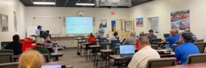 A classroom with several people seated at desks, a presenter at the front, and a presentation projected on a screen.