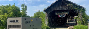 Industrial generator near a rustic covered bridge with a sign, set against a backdrop of trees and a partly cloudy sky.