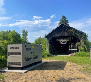 Industrial generator near a rustic covered bridge with a sign, set against a backdrop of trees and a partly cloudy sky.